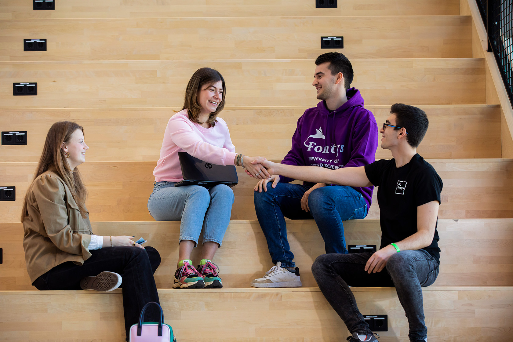 Vier (internationale) studenten zitten naast elkaar op een trap. Twee studenten schudden elkaar lachend een hand en de andere twee kijken hoe ze elkaar de hand schudden. 