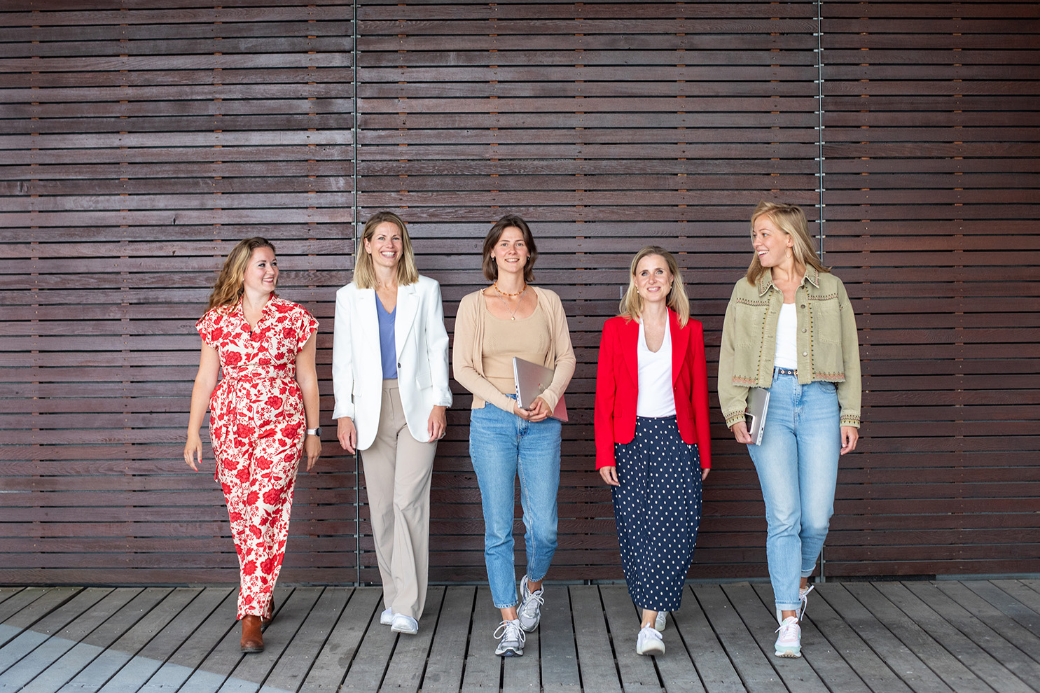 Vijf dames staan voor een muur. Dame links en dame rechts kijken lachend naar elkaar en de andere drie dames kijken lachend naar de camera. 