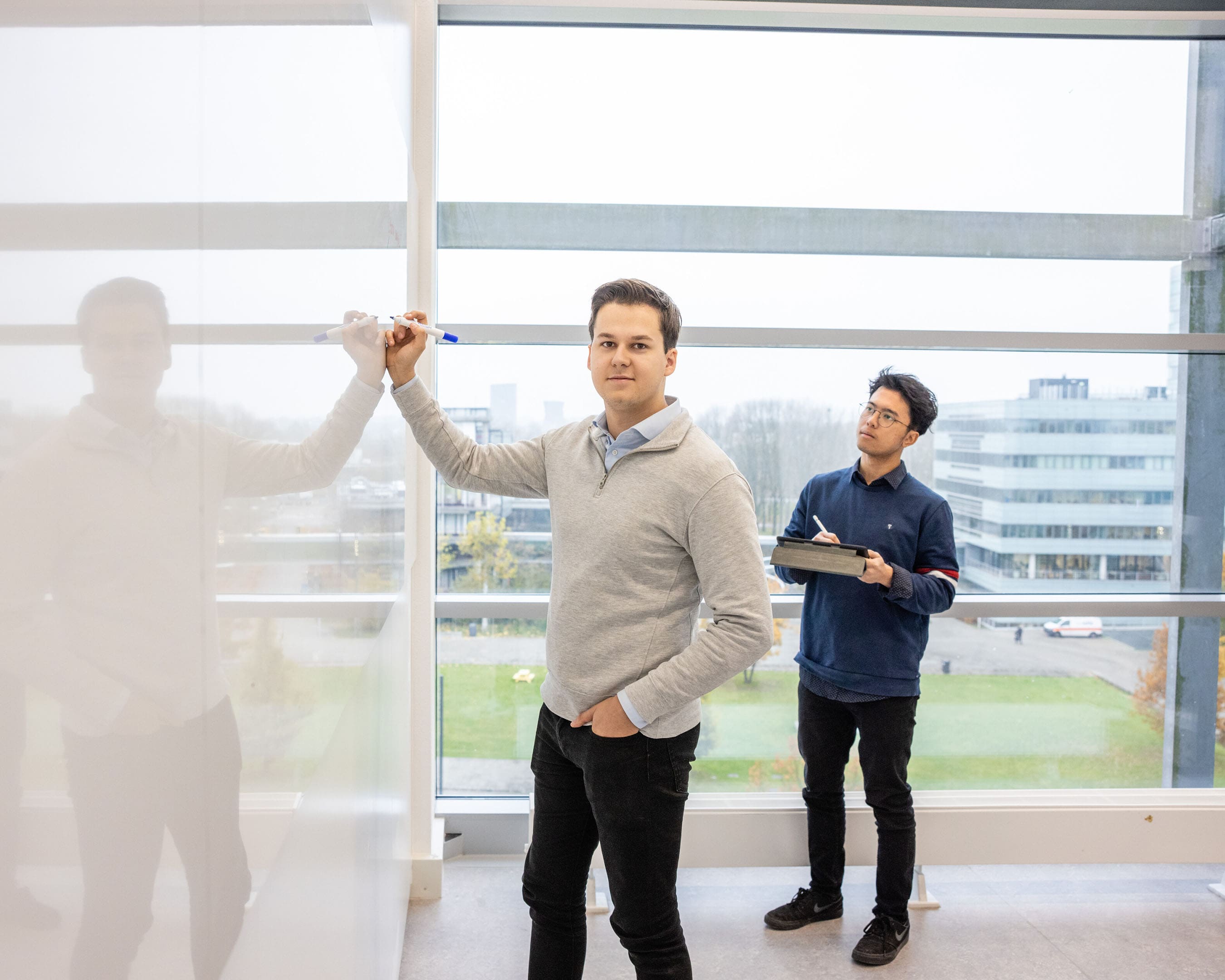 Twee studenten werken samen bij een whiteboard, met een uitzicht op gebouwen en groen.