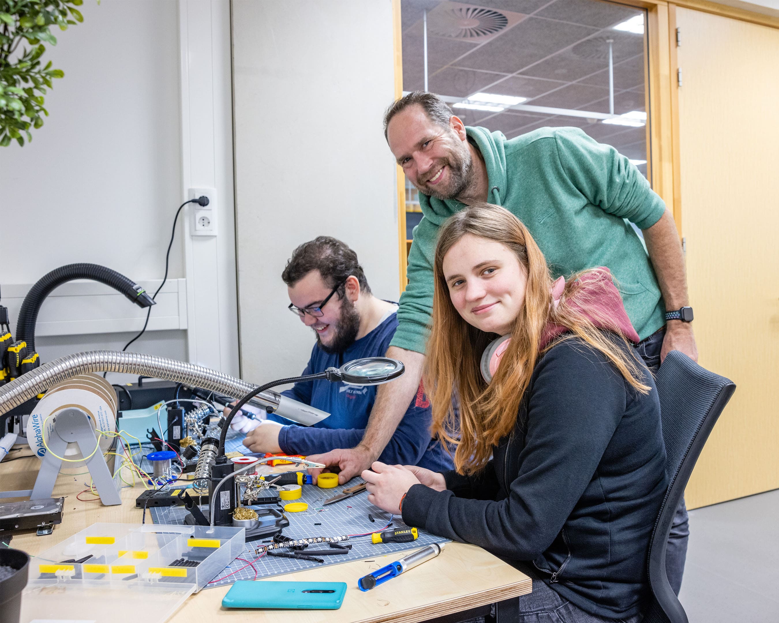 Studenten werken samen met docent aan technische projecten in een werkruimte, met gereedschap en elektronica op de tafel.