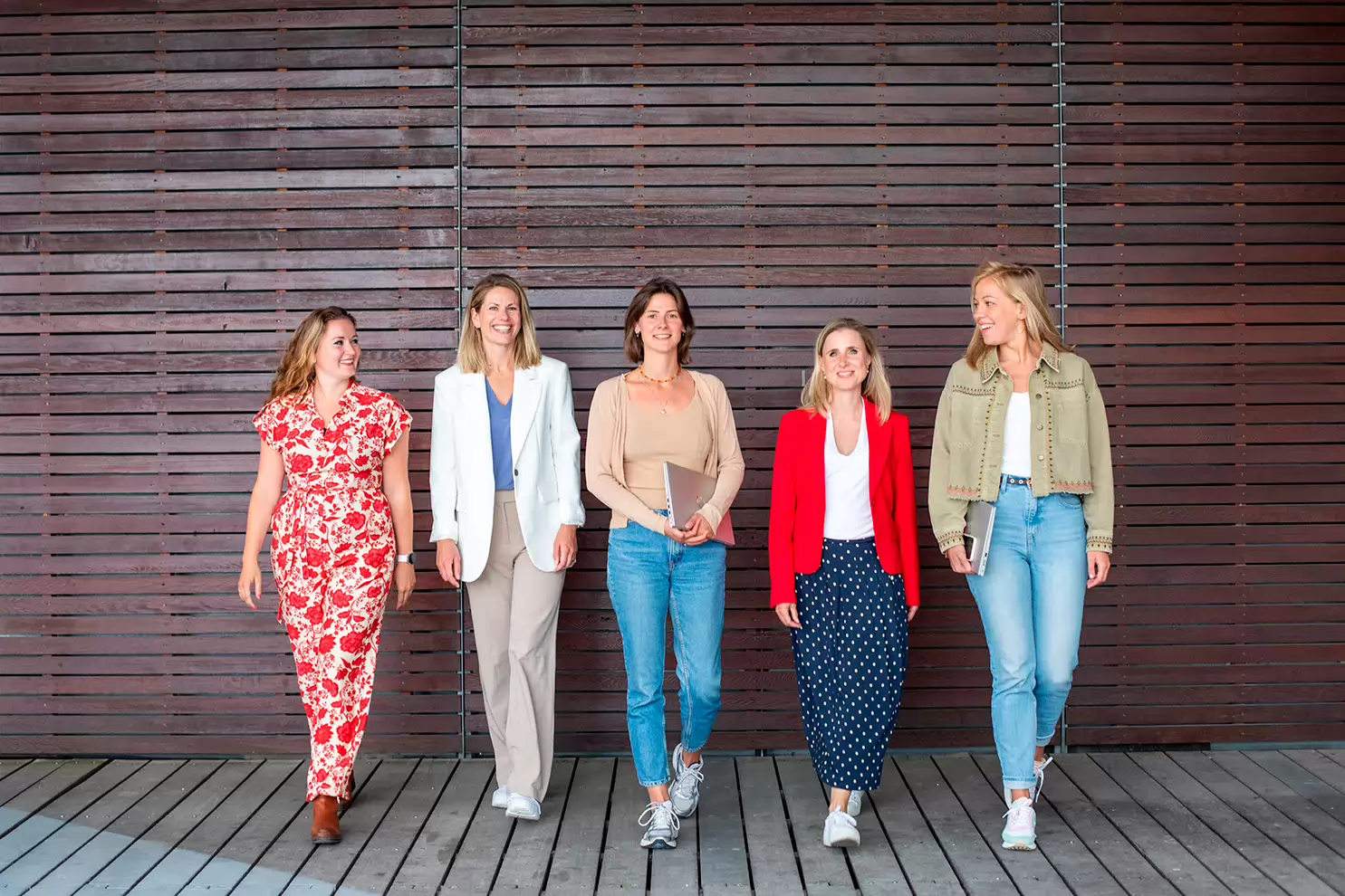 Vijf dames staan voor een muur. Dame links en dame rechts kijken lachend naar elkaar en de andere drie dames kijken lachend naar de camera.