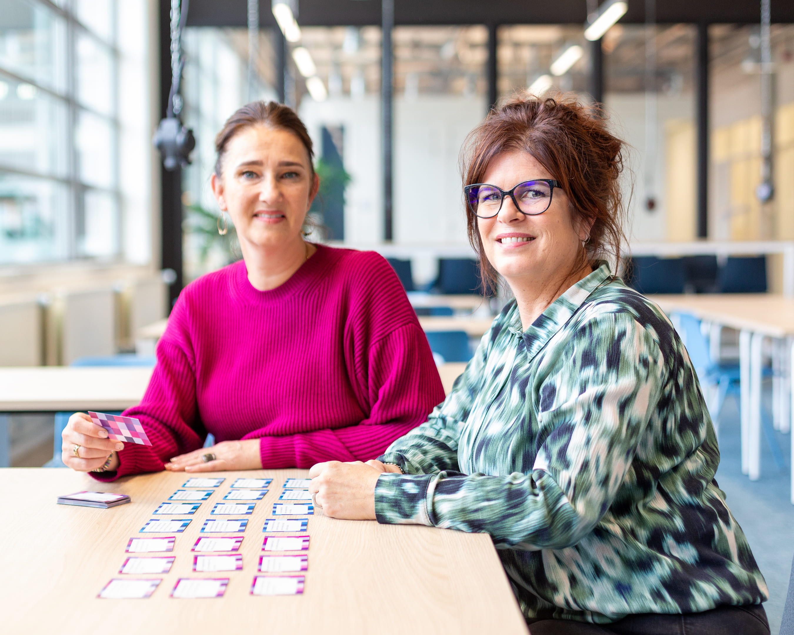 Twee studenten zitten aan een tafel en werken met kleurrijke kaartjes in een lichte, moderne ruimte.