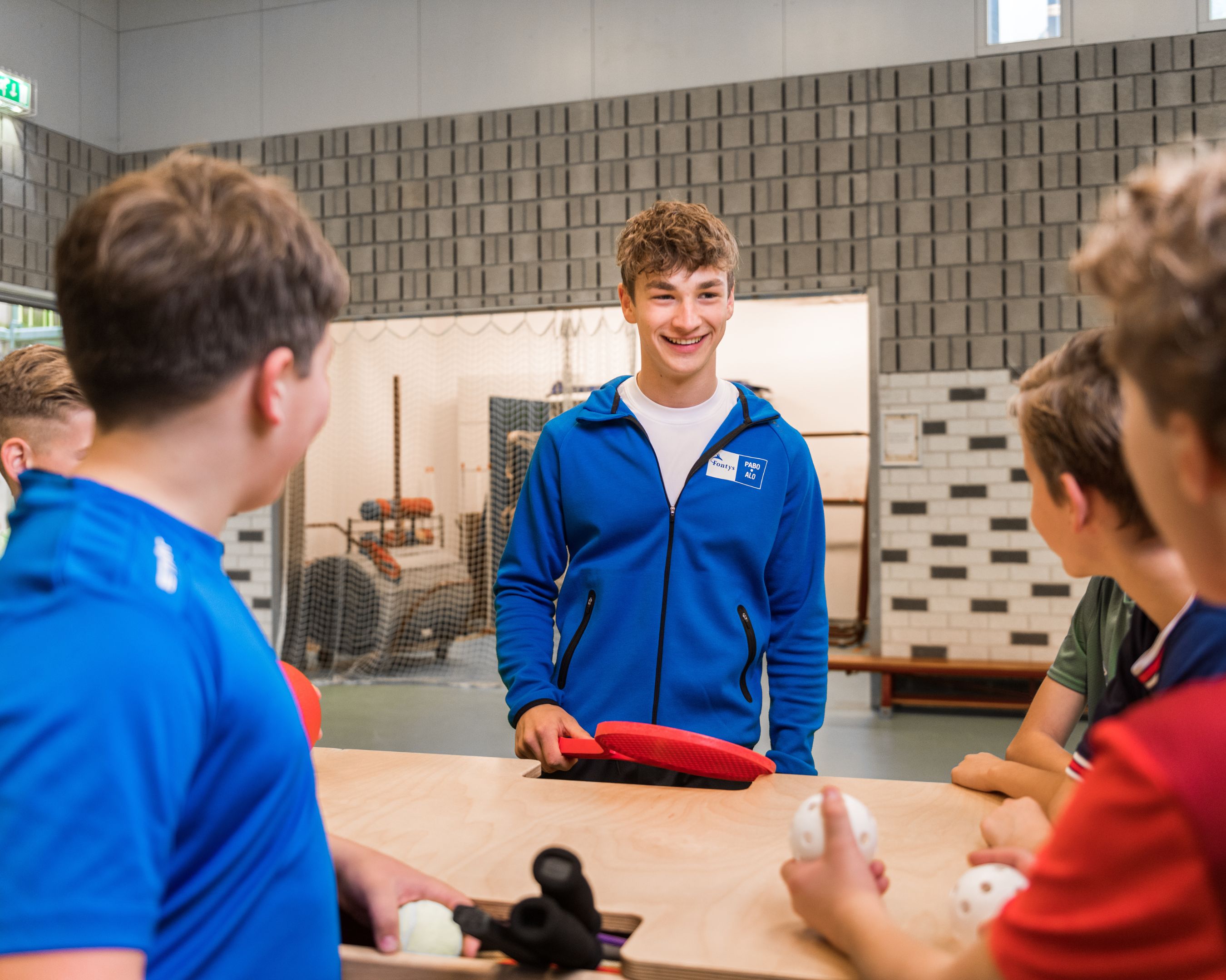 Studenten in sportkleren in een sportzaal kijken naar een tablet waarop instructies staan.