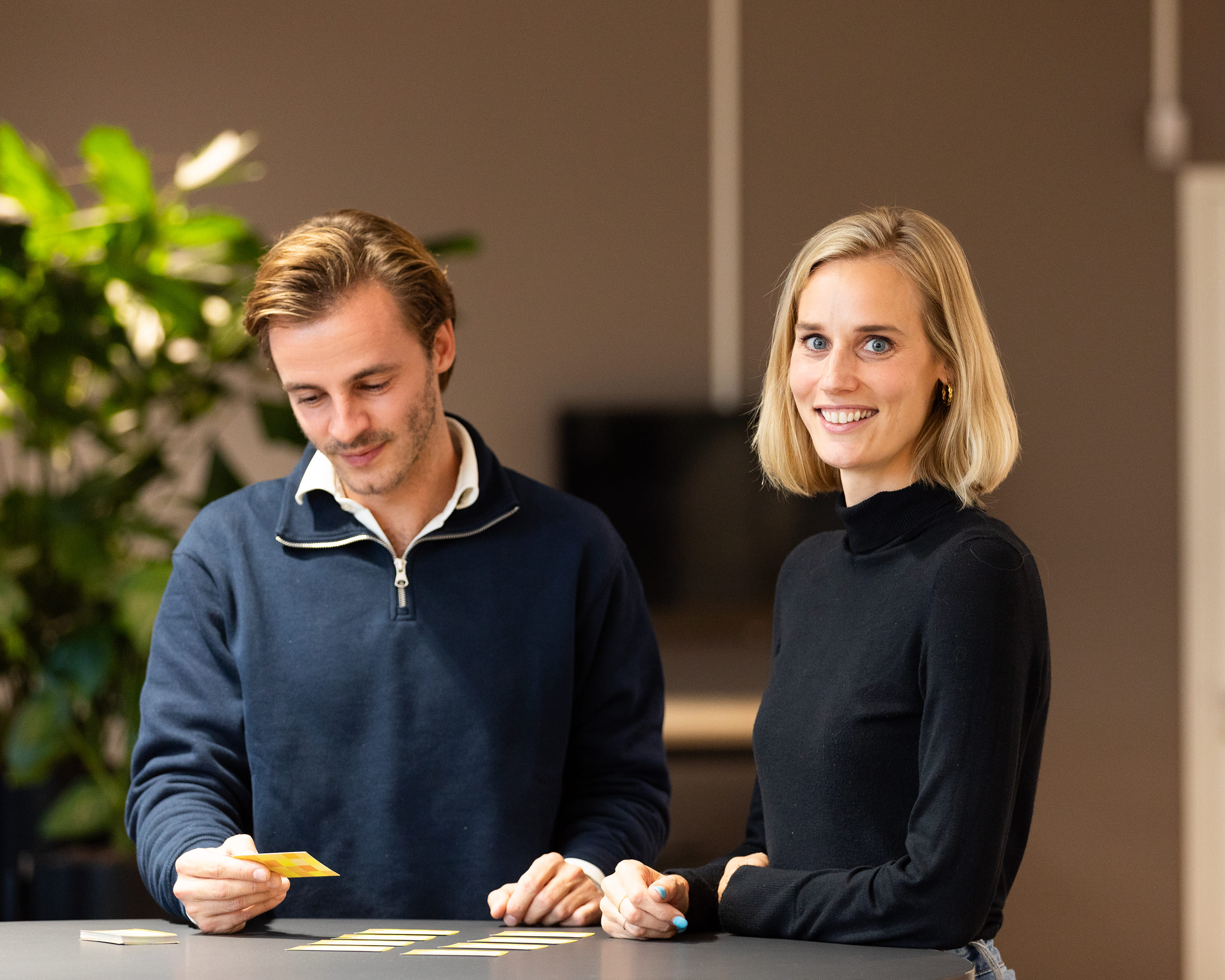 Twee studenten werken samen met kaartjes aan een tafel, terwijl een van hen glimlacht naar de camera.