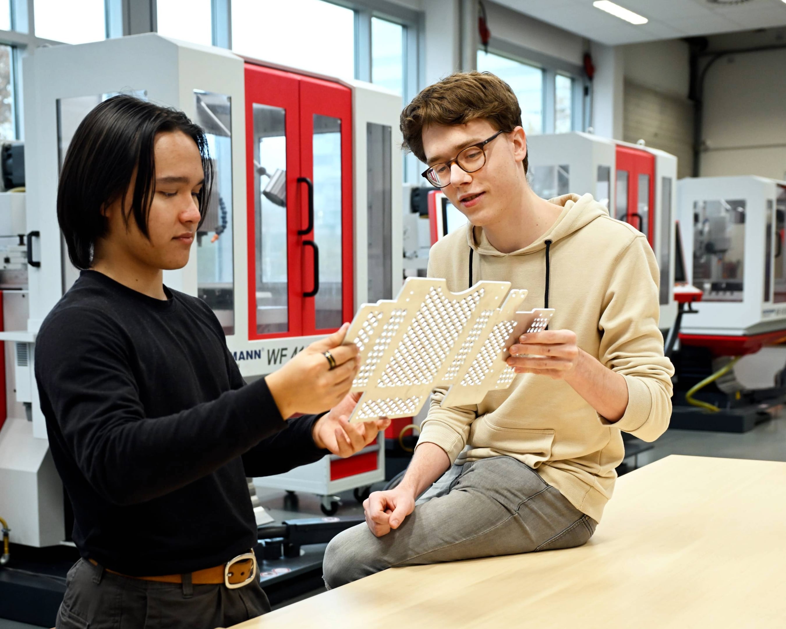 Twee studenten Werktuigbouwkunde bespreken technische onderdelen in een werkplaats, met machines op de achtergrond.