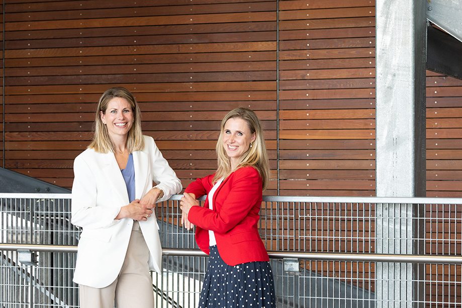 Twee vrouwen staan voor een muur en kijken lachend naar de camera. Zij gaan de samenwerking aan om trainingen en cursussen te bieden