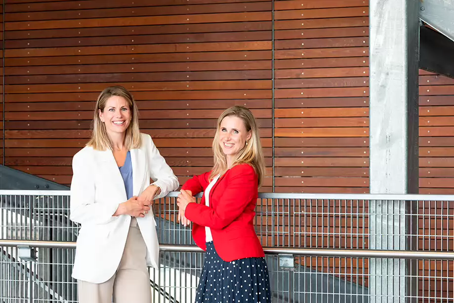 Twee vrouwen staan voor een muur en kijken lachend naar de camera. Zij gaan de samenwerking aan om trainingen en cursussen te bieden