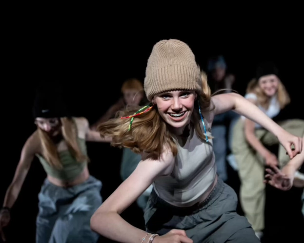 Jongeren dansen enthousiast op het podium, één meisje vooraan in actie.