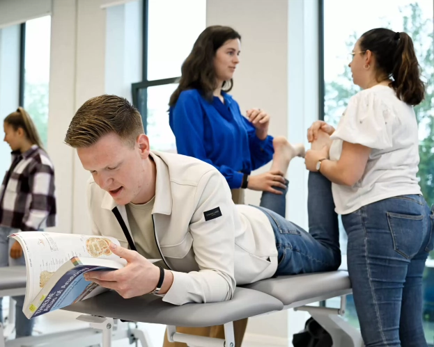 Fysiotherapiestudenten oefenen behandeltechnieken in een praktijkruimte, terwijl een student op een behandeltafel ligt en een studieboek leest.