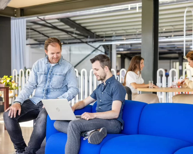 Twee studenten overleggen met een laptop op een blauwe bank in een moderne ruimte.