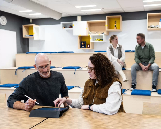 A student and a lecturer collaborate using a tablet, while two other people have a conversation in the background.