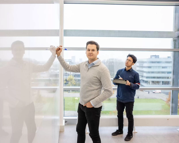 Twee studenten werken samen bij een whiteboard, met een uitzicht op gebouwen en groen.