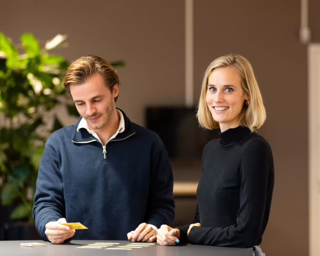 Twee studenten werken samen met kaartjes aan een tafel, terwijl een van hen glimlacht naar de camera.