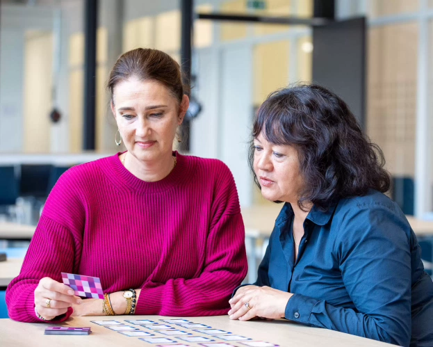 Twee personen werken samen aan een activiteit met kleurrijke kaartjes op een tafel in een lichte ruimte.