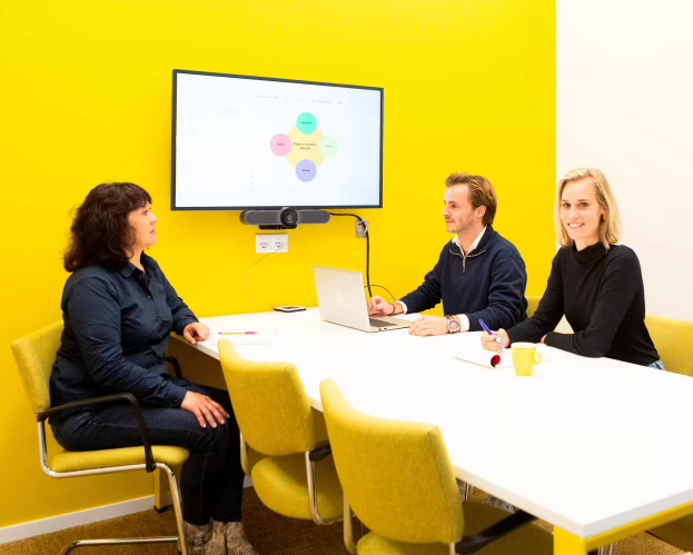 Drie personen in een vergaderruimte, werkend aan een project met een laptop en een presentatie op het scherm.