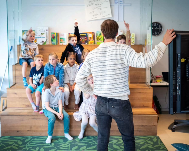 Docent voor de klas, waarbij de leerlingen in een theaterachtige opstellen luisteren en hun vinger opsteken.